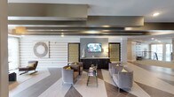 This lobby presents a sophisticated, contemporary design that sets a polished first impression. The space is anchored by a striking feature wall with a wall-mounted flat-screen television, flanked by two textured accent panels in warm gold and neutral tones, complemented by a dark wood credenza beneath. Architectural interest is added through layered ceiling details, including bold horizontal banded elements finished in contrasting tones of gray and white, adding depth and visual dimension overhead.

The flooring features a dynamic geometric pattern combining light wood-look planks with darker diamond-shaped tile insets, creating a visually engaging surface that defines the seating area. Plush gray upholstered lounge chairs with chrome legs are arranged symmetrically around a sleek glass-top coffee table, each accented with vibrant yellow throw pillows that introduce a lively pop of color.

Wall décor includes a large woven circular art piece, adding a natural texture contrast to the otherwise refined palette. Warm ambient lighting from table lamps enhances the inviting atmosphere. Overall, the lobby balances modern elegance with comfort, creating a welcoming and functional communal gathering area.