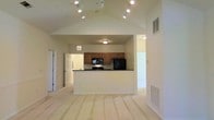 This single-story space is defined by its open-concept design and vaulted ceiling, which rises to a peak above the main living area, creating a sense of spaciousness and volume. Track lighting is mounted along the ceiling's ridge, providing versatile and well-distributed illumination throughout the space. The light-toned flooring extends across the living area, contributing to a bright and airy atmosphere.

The kitchen is positioned centrally, separated from the living area by a peninsula with a dark countertop that offers both prep space and a natural visual divide. Medium-toned wood cabinetry lines the kitchen walls, providing ample storage. Black appliances, including a built-in microwave and refrigerator, complement the dark countertops. The kitchen area features a lower, flat ceiling compared to the vaulted living space, subtly delineating the two zones.

Doorways visible from the main area suggest access to additional rooms beyond. Floor-level HVAC vents are positioned throughout, and the neutral wall tones keep the overall aesthetic clean and understated, making the space highly adaptable to various interior styles.