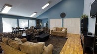 This single-floor space offers a warm and functional layout designed for comfortable gathering and relaxation. The main living area is anchored by vaulted ceilings that lend an airy, open feel, while bold blue accent walls provide a striking visual contrast against the crisp white trim and doors. The room is generously furnished with multiple seating arrangements, including plush sofas and patterned armchairs grouped around a dark wood coffee table, creating an inviting atmosphere for socializing or lounging. A large area rug defines the central seating zone, while hardwood-style flooring extends throughout, adding warmth and continuity to the space. Overhead, flush-mounted fluorescent light fixtures ensure the room is well-lit, complementing the natural light that filters in through multiple horizontal blinds-covered windows along the perimeter. Adjacent to the main seating area, a dining section features a dark-toned table with matching chairs, making the space versatile for both casual and more structured use. Two sets of double closet doors along one wall offer practical storage solutions. Decorative touches, including a dark medallion wall accent and potted greenery, add personality and a lived-in charm. Overall, the space balances practicality with comfort in a clean, organized setting.