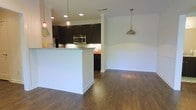 This single-floor space showcases an open-concept layout that seamlessly connects the kitchen, dining, and living areas, creating a cohesive and functional environment. The kitchen is a standout feature, outfitted with rich dark cabinetry that contrasts beautifully against light granite countertops and crisp white walls. Stainless steel appliances, including a built-in microwave, add a modern touch, while under-cabinet lighting ensures a well-lit workspace. A peninsula with a clean white finish defines the boundary between the kitchen and the adjoining areas, offering additional counter space and a natural gathering spot. Stylish glass and metal pendant lights hang above both the kitchen and the adjacent dining area, providing warm, focused illumination and serving as eye-catching design elements. A decorative column with classic detailing adds architectural interest and further delineates the open floor plan. Durable dark hardwood-style flooring runs throughout, grounding the space with warmth and continuity. The dining area is bathed in a clean, neutral palette with white wainscoting detail along the lower walls, lending a polished, timeless character. Recessed lighting complements the pendant fixtures, ensuring the entire space is bright and inviting throughout the day.