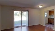 This single-floor space offers a clean, straightforward layout with a focus on functionality and natural light. The main living area features warm hardwood flooring that extends throughout, complementing the neutral white walls and white trim that create a bright, open atmosphere. A large sliding glass door serves as a prominent architectural feature, flooding the room with natural light while providing direct views of a well-maintained grassy outdoor area surrounded by mature trees. The sliding door also includes sheer curtains, allowing for adjustable privacy and light control. Ceiling-mounted flush lighting provides practical illumination for the space. A visible HVAC vent in the ceiling indicates a centralized climate control system. The white baseboards and clean wall finishes contribute to the overall tidy, move-in-ready appearance of the space. The combination of hardwood floors, abundant natural light, and outdoor access gives this space an open, airy feel that would suit a variety of uses. The simplicity of the design allows for versatile furnishing arrangements, making it adaptable to different living or working needs.