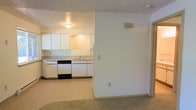 This single-floor space offers a straightforward, functional layout with a clear distinction between the living and kitchen areas. The carpeted living room is bright and open, featuring neutral white walls, a textured ceiling, and standard electrical outlets throughout. A half-wall partition separates the living space from the kitchen, creating a defined yet connected floor plan.

The kitchen is equipped with white cabinetry that spans both upper and lower sections, providing ample storage. A dishwasher and refrigerator are included among the appliances, complemented by a countertop sink positioned beneath overhead cabinets. The kitchen is finished with tile or hard flooring, contrasting with the carpet in the adjoining living area. A ceiling-mounted light fixture illuminates the kitchen, maintaining a clean and practical atmosphere throughout.

Overall, the space presents a simple, neutral palette with white walls and light-toned finishes, making it a versatile and easy-to-customize environment. The open sightlines between the main living and kitchen areas contribute to a cohesive feel across the single-floor layout.