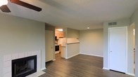 This single-floor space offers a practical and functional layout centered around an open-concept living area. The living room is a generous, rectangular space finished with dark hardwood-style flooring and neutral sage-green walls, creating a calm, understated atmosphere. A fireplace with a simple white surround anchors one wall, adding a cozy focal point to the room. The textured popcorn ceiling runs throughout, and a partial ceiling beam is visible near the entry, adding a subtle architectural detail.

The kitchen is partially separated from the living area by a half-wall pass-through counter, which maintains an open feel while defining the two spaces. Inside the kitchen, light wood cabinetry lines the walls, complemented by under-cabinet lighting that illuminates the workspace. A white refrigerator and a range with a black oven are visible, along with a range hood above. A door within the kitchen provides additional access, likely to a utility area or exterior.

White baseboards trim the perimeter throughout, providing clean contrast against the flooring. The overall design is modest and move-in ready, with a neutral palette that allows for flexible furnishing and personalization.