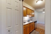 Located at 39670 Greenview Pl in Plymouth, Michigan, this two-bedroom, one-bathroom residence offers a well-organized single-floor layout. The galley-style kitchen is a functional highlight, featuring warm honey-toned wood cabinetry, granite-look countertops, stainless steel appliances including a dishwasher and refrigerator, and hardwood-style flooring. Ample upper and lower cabinet storage lines both sides of the kitchen, maximizing utility in the space. The entrance leads conveniently into the kitchen and living areas, with a hallway connecting the rest of the home. The generously sized primary bedroom is carpeted and bathed in natural light through a wide, multi-panel window overlooking a green outdoor landscape. The second bedroom is also carpeted and includes a closet, offering a bright and neutral space. The full bathroom features a wood-framed vanity with granite-look countertops, a single sink, tile surround, and a shower-tub combination. Throughout the home, neutral wall tones and light-colored carpeting create a clean, understated aesthetic. The layout, as seen in the dollhouse view, is efficient and well-proportioned, making the most of the available floor space.