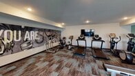This fitness center in Saint Cloud, Minnesota, offers a comprehensive workout environment designed to accommodate a wide range of exercise needs. The space is laid out across a single floor and is organized into distinct workout zones, each outfitted with purposeful equipment and thoughtful design details.

The cardio area features multiple treadmills and stationary bikes positioned in front of a wall-mounted flat-screen television, making extended sessions more engaging. A large, artistic mural depicting fitness-themed imagery serves as a bold focal point in this zone, reinforcing the space's energetic atmosphere.

The strength training section includes a multi-station cable machine, a flat weight bench, and a well-stocked dumbbell rack with floor-to-ceiling mirrors that visually expand the space. A rowing machine and additional free weights round out the functional fitness offerings.

The flooring throughout consists of mixed-pattern, multi-toned carpet tiles in earthy and teal hues, adding visual texture underfoot. Teal and neutral-toned accent walls contribute to a cohesive color palette. Recessed lighting illuminates the space evenly, while large windows along one wall introduce natural light.

Practical amenities include a water fountain station and a dedicated corner for stability balls and medicine balls. A motivational phrase — "The Stronger You Are, The Better You Feel" — is displayed prominently across one wall, reinforcing the space's encouraging, fitness-forward character.