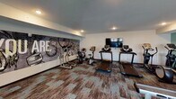 This fitness center in Saint Cloud, Minnesota, offers a comprehensive workout environment designed to accommodate a wide range of exercise needs. The space is laid out across a single floor and is organized into distinct workout zones, each outfitted with purposeful equipment and thoughtful design details.

The cardio area features multiple treadmills and stationary bikes positioned in front of a wall-mounted flat-screen television, making extended sessions more engaging. A large, artistic mural depicting fitness-themed imagery serves as a bold focal point in this zone, reinforcing the space's energetic atmosphere.

The strength training section includes a multi-station cable machine, a flat weight bench, and a well-stocked dumbbell rack with floor-to-ceiling mirrors that visually expand the space. A rowing machine and additional free weights round out the functional fitness offerings.

The flooring throughout consists of mixed-pattern, multi-toned carpet tiles in earthy and teal hues, adding visual texture underfoot. Teal and neutral-toned accent walls contribute to a cohesive color palette. Recessed lighting illuminates the space evenly, while large windows along one wall introduce natural light.

Practical amenities include a water fountain station and a dedicated corner for stability balls and medicine balls. A motivational phrase — "The Stronger You Are, The Better You Feel" — is displayed prominently across one wall, reinforcing the space's encouraging, fitness-forward character.