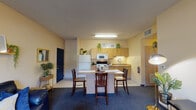 This single-floor unit is thoughtfully designed to accommodate shared living, offering multiple private bedrooms, a communal living area, a functional kitchen, and bathrooms. The open-concept kitchen and dining area serves as a central hub, outfitted with light wood cabinetry extending to the ceiling, a full suite of white appliances including a refrigerator, electric range, and over-the-range microwave, plus a countertop island with bar seating — well-suited for shared meal preparation and casual dining. Tile flooring runs through the kitchen, transitioning to plush carpet in the adjoining living area, where warm beige walls and a cozy seating arrangement create an inviting common space. Each private bedroom features carpeted floors, a window with blinds and curtain panels admitting natural light, a twin bed frame, dedicated desk workspace, and closet storage — providing residents with a functional personal retreat. The bathrooms are finished with tile flooring, a tub/shower combination, vanity with mirror, and ample storage. Consistent warm-toned walls and practical layouts throughout give the unit a cohesive, livable character.
