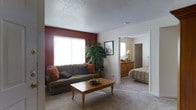 This single-floor residence offers a comfortable and well-appointed layout designed for easy living. The living room serves as the heart of the home, featuring a striking deep red accent wall that adds warmth and character to the space. A large window allows natural light to fill the room, highlighting the plush wall-to-wall carpeting underfoot. The textured ceiling adds a classic touch throughout. The living area provides ample room for seating and a central coffee table, creating a relaxed and inviting atmosphere. A tall indoor plant adds a refreshing natural element, complementing the warm color palette of earthy tones and neutral walls. An open doorway connects the living room directly to the bedroom, which is generously sized and features a wooden dresser, bedside lighting, and its own window that brings in additional natural light. The bedroom continues the carpeted flooring and neutral wall tones, creating a cohesive and calming aesthetic throughout the home. The overall ambiance is warm, lived-in, and functional, making this a practical and inviting single-floor living space well-suited for comfortable everyday life.