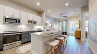 This single-floor residence offers a thoughtfully designed open-concept layout that seamlessly connects the kitchen, dining, and living areas. The kitchen is a standout feature, showcasing a two-tone cabinetry scheme with crisp white upper cabinets and slate-gray lower cabinets, a classic white subway tile backsplash with under-cabinet lighting, a large center island with bar seating, and a full suite of stainless steel appliances including a French door refrigerator, range, microwave, and dishwasher. Light wood-style flooring flows throughout the main living areas, enhancing the bright and airy atmosphere. The living room benefits from abundant natural light streaming through large windows dressed in patterned drapes, complemented by a ceiling fan overhead. A private door leads to an outdoor area, further extending the sense of openness. The primary bedroom is a calm retreat, featuring textured accent walls, large windows, and plush carpeting. Its en-suite bathroom is finished with dual-sink gray vanity cabinetry, a glass-enclosed walk-in shower, and light tile flooring. A spacious walk-in closet with built-in shelving and double hanging rods provides generous storage. The overall ambiance throughout the residence is warm, modern, and cohesive.