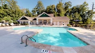 Located at 9525 US-78 Highway in Ladson, South Carolina, this single-story community facility centers around a generously sized outdoor swimming pool with crystal-clear blue water. The pool area is framed by a classic craftsman-style clubhouse building, characterized by green board-and-batten siding accents, a pitched roof with dormers, and wall-mounted lantern lighting, lending the space a welcoming residential charm.

The pool itself features multiple entry points, including stainless steel handrail ladders, with clearly marked depth indicators along its perimeter. A wide concrete pool deck surrounds the water, providing ample space for circulation and relaxation. The sun deck is well-equipped with multiple reclining lounge chairs, ideal for sunbathing, as well as a shaded seating area outfitted with patio tables, chairs, and a large umbrella for those seeking respite from the South Carolina sun.

The entire pool area is enclosed by a black wrought iron fence, ensuring safety and privacy while maintaining open sightlines to the lush, mature tree canopy that borders the property. The natural greenery creates a serene, park-like backdrop that enhances the overall outdoor ambiance. The clubhouse building adjacent to the pool appears to provide restroom and support facilities, making this a fully functional and inviting community gathering space.