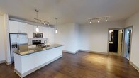 This single-floor space showcases a thoughtfully designed open-concept layout that seamlessly connects the kitchen and living area. The kitchen is outfitted with crisp white cabinetry, dark granite countertops, and stainless steel appliances, including a built-in microwave and electric range. A kitchen island with a sink serves as a functional centerpiece, offering additional workspace and a natural division between the kitchen and the expansive living area. Pendant lighting above the island adds a refined touch, complementing the track lighting installed throughout the main living space. Rich hardwood flooring runs continuously across both areas, grounding the space with warmth and visual continuity. The living area is generously proportioned, with clean white walls and neutral tones that create a bright, airy atmosphere. The overall design emphasizes a balance of modern aesthetics and practical functionality, making the space feel both polished and livable.