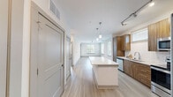 Located at 901 River Road in Charlottesville, Virginia, Unit 202 is a thoughtfully designed single-floor residence offering a comfortable and modern living experience. The open-concept kitchen and living area forms the heart of the unit, where light wood-tone flooring flows seamlessly throughout, creating a warm and cohesive aesthetic. The kitchen is outfitted with rich wood-grain cabinetry, white quartz countertops, a classic subway tile backsplash, and a full suite of stainless steel appliances including a range, over-the-range microwave, dishwasher, and refrigerator. A central kitchen island with a clean white finish provides additional prep and seating space, while pendant lighting adds a refined touch overhead. The living area is bright and open, benefiting from multiple windows dressed with horizontal blinds that invite natural light while maintaining privacy. Outdoor views of the surrounding greenery are visible from the living space, which also features access to an exterior door. A ceiling fan adds comfort to the room. The walk-in closet offers generous storage with wire shelving systems and carpeted flooring, providing practical organization space. Neutral wall tones and clean architectural lines throughout the unit contribute to a fresh, modern ambiance well-suited for comfortable everyday living.