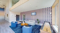 Located at 10 Patriot Pky in South Weymouth, MA, this single-floor lobby space offers a striking first impression with its contemporary architectural design and vibrant interior details. The layout is divided into two interconnected hallway areas that together create a welcoming and functional entry experience.

Soaring double-height ceilings define the main lounge area, amplifying the sense of openness and grandeur. Floor-to-ceiling windows along the perimeter flood the space with natural light while framing views of the surrounding streetscape and greenery. Warm wood accents on the upper wall panels complement the light-toned luxury vinyl plank flooring that runs throughout.

A bold, geometric-patterned feature wall in shades of red, blue, and white serves as the visual centerpiece of the lounge area, anchoring a curved sofa, coordinating blue armchairs, a round wood coffee table, and a vibrant blue area rug. A wall-mounted television and a shuffleboard table add recreational appeal. High-top tables with black bar stools are positioned near the large windows, offering casual seating with outdoor views.

Artwork displayed throughout — including travel-themed prints and patriotic graphic pieces — reinforces a curated, community-oriented aesthetic. A secondary hallway connects to secured entry doors, featuring clean white walls with a textured wainscoting detail, smooth flooring, and smart access hardware, completing this polished and purposeful shared space.