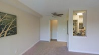 Located at 1701 Lee Rd. in Winter Park, Florida, this single-floor space offers a comfortable and functional layout designed for everyday living. The main living area is finished with wall-to-wall carpeting in a neutral tone, contributing to a warm and inviting atmosphere. A ceiling fan is centrally mounted beneath a textured ceiling, providing both air circulation and a subtle decorative element. The walls are painted in a soft, neutral beige palette, creating a clean and understated aesthetic throughout. A doorway from the living area leads to an adjacent hallway or secondary space, suggesting a well-connected floor plan. To one side, an open pass-through counter connects the living area to the kitchen, allowing for easy interaction between the two spaces. The kitchen features visible shelving and appliances, including what appears to be a washer and dryer unit, indicating in-unit laundry convenience. The overall layout is practical and efficiently organized, making good use of the available space. The neutral finishes and open sightlines lend the interior a bright, airy feel. Situated in the desirable Winter Park community, the property benefits from its central Florida location with access to local amenities, parks, and dining.
