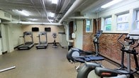 This fitness room offers a practical and functional workout environment housed within a space that blends industrial and historic character. The ceiling features exposed ductwork and piping painted white, lending an open, utilitarian feel, while fluorescent lighting ensures the space is well-lit throughout. One of the most distinctive architectural elements is the exposed brick wall along one side of the room, adding a rugged, historic charm that contrasts with the otherwise modern fitness equipment.

Natural light filters in through multiple windows positioned at street level, providing ventilation and a connection to the outdoors. The flooring consists of durable, rubberized material well-suited for exercise use. The room is outfitted with a selection of cardio equipment, including multiple treadmills, stationary bikes, and elliptical trainers from Life Fitness, a reputable fitness brand. Two wall-mounted flat-screen televisions are positioned for easy viewing during workouts.

Built-in cabinetry along one wall provides storage space. The overall layout is organized and spacious enough to accommodate multiple users simultaneously, making it a convenient and well-appointed fitness facility.