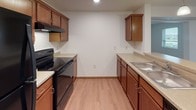 This single-floor residence offers a functional and comfortable layout across several distinct living spaces. The entry area flows into an open-concept zone where light hardwood-style flooring connects the kitchen and dining areas, creating a cohesive, welcoming atmosphere. The galley-style kitchen is well-equipped with warm medium-toned wood cabinetry, light countertops, a black electric range, a full-size black refrigerator, and a double sink — providing ample storage and prep space for everyday living. A peninsula counter with seating potential further enhances the kitchen's utility.

The living area features plush wall-to-wall carpeting and a ceiling fan with integrated lighting, lending a relaxed, comfortable feel. A sliding door provides access to the exterior, allowing natural light to fill the space. Two additional carpeted rooms serve as bedrooms, each with neutral white walls and access to natural light via windows with blinds. The full bathroom is finished with warm wood vanity cabinetry, a white countertop, a large mirror, tile flooring, and a full shower-tub combination. A dedicated in-unit laundry room houses a full-size top-loading washer and dryer with overhead wire shelving, adding significant day-to-day convenience. An electrical panel is also accessible within the unit.