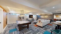 Located at 44 Hill St in Morristown, NJ, this well-appointed amenity space offers a thoughtfully designed single-floor layout encompassing a game room, living room, and exercise room. The game room serves as a centerpiece, featuring a full-size pool table illuminated by striking geometric pendant chandeliers. A wall-mounted cue rack and a textured cork accent wall add warmth and character, while clerestory windows allow natural light to filter in. Adjacent to the game room, a shuffleboard table is tucked against a dramatic stacked-stone accent wall, adding another layer of entertainment. The living room area is furnished with a generous sectional sofa, modern coffee table, and additional seating — all anchored by a patterned multi-tone carpet that runs throughout the space. A wall-mounted flat-screen TV, a compact bar-height counter, and pendant lighting contribute to the lounge's relaxed yet refined atmosphere. Crown molding and recessed lighting elevate the overall aesthetic. The exercise room completes the layout, providing a dedicated space for fitness. Throughout, the design palette blends neutral tones with vibrant orange accent rugs and teal details, creating a cohesive, contemporary feel ideal for recreation and socializing.