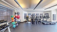 This fitness center is designed to support a comprehensive workout experience within a single, open-plan floor. The space is divided into functional zones, each dedicated to a different category of exercise. The cardio area is outfitted with multiple treadmills, elliptical trainers, and a stationary bike, all positioned near a row of windows that allow natural light to filter in. A large wall mirror on one side amplifies the sense of space and assists with form during workouts.

The strength-training section features a multi-function cable machine, a weight bench, and a full rack of dumbbells neatly organized for easy access. A wall-mounted flat-screen television provides entertainment throughout the space. On the opposite side, a colorful rack of medicine balls and stability balls in various sizes adds a functional training element to the room.

The flooring throughout is durable rubberized material suited for high-impact activity. Recessed and cove lighting in the ceiling creates a well-lit, energizing atmosphere. A standout feature is the glass sliding door at one end, which opens directly to an outdoor pool area, offering both refreshing views and convenient access. A vending machine is also available near the exit for post-workout refreshments.