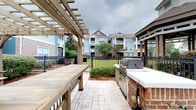 Located at 1001 Bear Island Rd in Summerville, South Carolina, this outdoor amenity space is designed for community gathering and al fresco entertaining. The patio is anchored by a wood-framed pergola that provides partial shade over a large communal dining table, creating an inviting setting for group meals and social events. Adjacent to the pergola, a brick-built outdoor kitchen station is equipped with a stainless steel multi-burner gas grill, offering a well-appointed setup for outdoor cooking. The patio surface features decorative stamped concrete pavers that add a refined aesthetic to the space. Wrought iron fencing with a classic design defines the perimeter, providing both structure and an open sightline to the surrounding green landscape. Manicured shrubbery and mature trees line the grounds, enhancing the natural ambiance of the area. In the background, multi-story residential buildings with blue and neutral-toned exteriors frame the community setting, reflecting the well-maintained character of the neighborhood. Overall, this outdoor space combines functional amenities with an approachable, community-focused design ideal for residents seeking outdoor leisure and entertainment options.