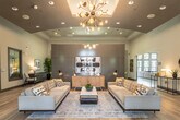 This single-floor amenity and leasing center is thoughtfully designed to blend functionality with contemporary style. The main reception area greets visitors with soaring ceilings, warm light wood flooring, and an open, airy layout. Workstations and seating are strategically arranged to accommodate both staff and guests, while large arched windows and glass-paneled entry doors flood the space with natural light and offer views of the surrounding greenery.

Adjacent to the reception zone, a comfortable lounge area features a tufted upholstered sofa, layered area rugs, curated wall art, and a flat-screen display — creating an inviting space for relaxation or informal meetings. A tray ceiling with recessed lighting adds architectural depth and a refined ambiance to this section.

A dining lounge transitions seamlessly from the main areas, furnished with a mix of round and rectangular tables paired with vibrant yellow chairs. Floor-to-ceiling sheer curtains frame large windows and double glass doors that open to an outdoor patio, allowing natural light to filter softly throughout. Beyond the dining area, an open-plan gathering room with large-format tile flooring, a dining table, lounge seating, and a built-in bar area provides a versatile space for community events and socializing. Lush potted plants are placed throughout, adding a fresh, natural element to the overall design.