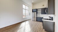 Located at 2000 North 2nd Street in Philadelphia's vibrant Northern Liberties neighborhood, this one-bedroom, two-bathroom residence spans multiple floors and offers a thoughtfully designed living experience. The main living level impresses with soaring ceilings and large, multi-pane windows that flood the space with natural light while framing dynamic city views. Light-toned hardwood-style flooring runs throughout, complementing the clean, modern aesthetic. The kitchen is well-appointed with sleek dark cabinetry, stainless steel appliances including a refrigerator, range, and microwave, white countertops, and open shelving — all combining function with contemporary style. The bedroom features floor-to-ceiling windows on two walls, offering sweeping views of the surrounding Philadelphia streetscape and filling the room with abundant daylight. In-unit laundry is tucked conveniently within the hallway area. The rooftop level is a standout feature, offering a wood-decked outdoor space with a green roof surround and panoramic views of the Philadelphia skyline — an exceptional retreat above the city. Multiple staircases connect each level seamlessly, adding to the multi-floor character of this urban residence.
