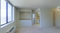 This single-floor space presents a straightforward, functional layout designed for comfortable everyday living. The main living area is finished with light-toned carpeting that extends throughout, contributing to a clean, neutral aesthetic. A textured popcorn ceiling adds a classic touch overhead, while the white walls keep the interior bright and airy.

A distinctive built-in wall nook with an illuminated interior shelf serves as a focal point in the living area, offering both display and storage potential. Beneath the nook, a wooden countertop ledge provides additional surface space, making it a practical feature for everyday use.

An open passageway connects the living area to the kitchen, which is outfitted with white cabinetry on both upper and lower levels, offering ample storage. The kitchen features light-colored countertops and tile flooring, maintaining the consistent neutral palette found throughout the space. A hallway leads further into the unit, suggesting additional rooms beyond what is immediately visible. A wall-mounted heating unit is positioned along the lower wall of the living area, ensuring year-round comfort. Overall, the space offers a clean, well-organized environment with practical built-in features.