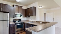 Located at 1000 Broadstone Way in Apex, NC, this single-floor residence offers a thoughtfully designed layout with functional living spaces throughout. The kitchen is a standout feature, equipped with stainless steel appliances including a refrigerator, range, and microwave, all complemented by rich dark-stained cabinetry, granite-style countertops, and a peninsula that provides additional workspace and bar-style seating. Durable wood-look flooring grounds the kitchen area, adding warmth and continuity. The open living area flows naturally from the kitchen, featuring plush carpeting, neutral-toned walls, and a vaulted ceiling with a ceiling fan, creating an airy and comfortable atmosphere. Large windows and a sliding glass door invite abundant natural light and offer views of the surrounding greenery. The bedroom is generously sized with soft carpeting, a ceiling fan with light fixture, a bright window with outdoor views, and convenient access to a walk-in closet. The bathroom is well-appointed with a dark-stained vanity, granite-style countertop, tile flooring, and a shower-tub combination. Additional storage is provided throughout, contributing to the overall practicality of this well-maintained Apex residence.