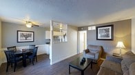 This single-floor space offers an open-concept layout that seamlessly connects the living, dining, and kitchen areas into one cohesive environment. The living area is anchored by a comfortable seating arrangement centered around a dark coffee table, creating a relaxed and inviting atmosphere. A ceiling fan overhead provides both light and airflow to the space.

The kitchen is partially separated by a half-wall partition featuring a pass-through opening with a built-in shelf, allowing natural sightlines between the cooking and living spaces while maintaining a defined boundary. White cabinetry lines the kitchen walls, contributing to a clean, bright aesthetic in that area.

Adjacent to the kitchen, a dining nook accommodates a small table and chairs, making it well-suited for everyday meals. The rich, dark wood-style flooring runs consistently throughout, providing a warm contrast to the neutral gray walls and bright white trim and baseboards.

The textured popcorn ceiling adds a classic residential character, while framed artwork on the walls enhances the overall décor. A hallway door visible from the main living space suggests access to additional private rooms beyond the open-plan area.