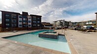 This outdoor amenity space is designed to serve as a central gathering hub for its residential community. At the heart of the area sits a generously sized swimming pool with crystal-clear blue water, complemented by an adjacent raised spa, both enclosed within a secure black iron fence that defines the perimeter of the courtyard. The surrounding pool deck is finished in large-format concrete pavers, providing ample space for sunbathing and relaxation, with reclining lounge chairs and side tables thoughtfully arranged throughout.

A standout feature of the space is the substantial brick outdoor fireplace, which anchors a dedicated seating area furnished with vibrant red cushioned chairs and benches. This zone creates an inviting, social atmosphere suited for year-round use. Adjacent to the fireplace lounge is a clubhouse structure clad in sage green metal siding with expansive windows, offering views into what appears to be an interior fitness or amenity room. Additional bar-height seating and casual furniture are positioned near this building, extending the outdoor living experience.

The surrounding multi-story residential buildings, finished in a combination of dark brown, green, and neutral tones, frame the courtyard and provide an urban backdrop. The open-sky setting allows abundant natural light to fill the space throughout the day, enhancing its welcoming and functional character.