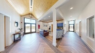This single-story community facility spans a generously sized footprint, thoughtfully designed to balance functionality with a warm, welcoming aesthetic. The entry lobby immediately impresses with soaring vaulted ceilings clad in warm wood planking, a statement pendant light fixture, and large glass double doors that flood the space with natural light and offer views of the surrounding exterior. The herringbone-patterned wood-look tile flooring flows throughout the main corridors and common areas, lending a cohesive and polished character to the layout.

The combined kitchen and living area features bold navy-blue cabinetry with decorative tile backsplash, creating a vibrant focal point. Adjacent hallways, adorned with curated artwork, connect seamlessly to additional rooms. The dedicated living room showcases elevated ceilings with exposed wood beam accents, built-in shelving, wall-mounted television, and comfortable leather seating arranged around a patterned area rug. A separate, secondary living area offers a tray ceiling, ceiling fan, multiple windows, and a dining space.

The exercise room is well-equipped with cardio machines, free weights, and a cable system, benefiting from direct outdoor access and natural light. The exterior features board-and-batten siding with stacked stone accents and a covered patio.