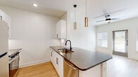 This thoughtfully designed 1-bedroom, 2-bathroom residence offers a clean, contemporary aesthetic across a single floor. The open-concept kitchen and living area serves as the heart of the home, anchored by a peninsula with dark stone countertops, a matte black undermount sink, and stainless steel appliances including a refrigerator, range, and microwave. White shaker-style cabinetry with black hardware lines the kitchen walls, complemented by a built-in wine rack and subway tile backsplash. Elegant gold-toned pendant lights hang above the peninsula, adding a stylish contrast to the neutral palette. Light hardwood flooring flows seamlessly throughout the entire residence, enhancing the sense of openness and continuity. The living area receives ample natural light through windows and a glass-paneled exterior door, with a ceiling fan overhead. The bedroom is a well-proportioned retreat featuring a large window with blinds, a ceiling fan, and clean white walls. The primary bathroom offers a dark stone-topped vanity with white cabinetry, a framed mirror, and direct access to a walk-in closet with wire shelving. A second bathroom provides additional convenience. The overall ambiance is bright, minimal, and modern.