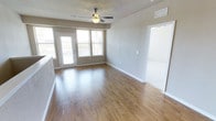 This single-floor space presents a well-lit, open living area defined by warm hardwood-style flooring that runs throughout the main room, lending a clean and cohesive aesthetic. A ceiling fan with a built-in light fixture is centrally mounted on the textured ceiling, providing both air circulation and ambient lighting. Natural light fills the space generously through a set of large windows fitted with horizontal blinds and a glass-paneled door, which appears to lead to an outdoor area, offering views of the exterior surroundings. The neutral, greige-toned walls complement the light wood flooring, creating a bright and versatile atmosphere. White baseboards frame the room cleanly, and an HVAC vent is visible near the ceiling, indicating a built-in climate control system. A doorway on the right side of the main room leads to an adjacent area with carpeted flooring, suggesting a separate room such as a bedroom. The overall layout feels open and functional, with a straightforward, contemporary design that provides a neutral canvas suitable for a variety of furnishing styles.