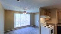 This single-floor residential space offers a functional and comfortable layout designed for everyday living. The main living area is bright and inviting, anchored by a large picture window fitted with vertical blinds that allows generous natural light to filter in while providing a view of the surrounding greenery. A ceiling fan with integrated lighting is centrally mounted on the textured ceiling, promoting air circulation and adding a practical touch to the room. Soft carpeting covers the living area floor, contributing to a warm and cozy atmosphere. Adjacent to the living space, the kitchen is efficiently designed and features a white electric range with overhead ventilation, warm-toned wood cabinetry providing ample storage, and a layout that keeps everything within easy reach. The neutral wall tones throughout create a cohesive and understated aesthetic that lends itself well to personalization. A white interior door near the kitchen offers access to additional areas of the unit. Overall, the space presents a straightforward, well-organized floor plan that balances the living and cooking areas in a compact yet livable arrangement.