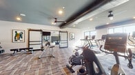 Located at 2074 Fox Valley Center in Aurora, IL, this well-appointed fitness center offers a comprehensive, multi-zone exercise environment designed to support a wide variety of workout routines. The main cardio area is generously sized, featuring a lineup of treadmills, elliptical trainers, a stair climber, and Matrix-brand cycling bikes, all bathed in natural light from large windows that offer views of the surrounding area. Overhead, exposed ductwork and ceiling fans lend an industrial-modern aesthetic, complemented by recessed lighting and a neutral, plaid-patterned carpet throughout. A dedicated free weights zone houses a full rack of dumbbells alongside adjustable benches and a cable resistance machine, providing ample space for strength training. Stylish barn-style glass doors connect to an additional flex area equipped with spinning bikes, a rowing machine, and a plyo box, offering space for functional and high-intensity workouts. A separate, carpeted studio room with full-length mirrors and wall-mounted TRX suspension trainers and stability balls provides an ideal setting for yoga, stretching, or group classes. The overall design strikes a balance between industrial character and contemporary cleanliness, creating an energizing and functional environment for fitness.
