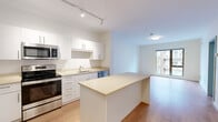 Located at 211 Rantoul Street in Beverly, MA, this thoughtfully designed one-bedroom, one-bathroom residence offers a clean, modern aesthetic across a single floor. The open-concept kitchen and living area serves as the heart of the unit, featuring crisp white cabinetry, light-toned countertops, a central island, and a full suite of stainless steel appliances including a range, over-the-range microwave, dishwasher, and refrigerator. Track lighting illuminates the kitchen, while the living area benefits from a large window that draws in natural light and provides views of the surrounding neighborhood. Light wood flooring runs throughout the unit, contributing to a warm, cohesive feel. The bedroom is a well-proportioned retreat with additional large windows that bring in ample natural light and offer outdoor views. Soft, neutral walls and the same wood flooring continue into this space, lending a calm and airy atmosphere. The bathroom is finished with a wood-toned vanity, white countertop, undermount sink, and neutral tile flooring. A dedicated laundry room houses a stacked washer and dryer unit with a wall-mounted storage shelf, adding everyday convenience. The unit's bright, minimalist design and functional layout make it a practical and comfortable residence in Beverly.