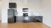 This single-floor space showcases a clean, contemporary design with a cohesive aesthetic carried throughout. The open-concept layout seamlessly connects the kitchen and living area, creating a spacious and airy feel enhanced by crisp white walls and warm-toned light wood flooring underfoot.

The kitchen is a standout feature, outfitted with a striking two-tone cabinet scheme — dark lower cabinets paired with white upper cabinets that extend toward the ceiling, maximizing storage. A white countertop and backsplash provide a bright contrast, while black appliances including a refrigerator, electric range, built-in microwave, and dishwasher lend a bold, modern edge. A stainless steel sink completes the functional workspace.

The open living area is generously proportioned, bathed in natural light from a large window along one wall. The unobstructed layout offers exceptional flexibility for furniture arrangement. Recessed lighting provides additional illumination throughout.

The bathroom maintains the space's modern sensibility with light wood-look flooring, a white tile surround enclosing a full soaking tub and shower, a floating vanity with an integrated sink, and a large mirror. The clean, minimal palette ties the bathroom seamlessly to the overall design language of the space.