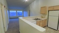 This single-floor space is defined by its striking vaulted ceilings that lend an airy, expansive quality to the open-concept layout. The kitchen is positioned at the heart of the home, anchored by a wraparound peninsula that provides generous counter space finished in light-toned laminate. Light wood cabinetry lines both the upper and lower sections, offering ample storage, while a double-basin stainless steel sink sits conveniently along the peninsula. The kitchen flows seamlessly into the living area, which is finished with wall-to-wall carpeting that adds warmth underfoot. A large window at the far end of the living space frames a natural wooded view, filling the room with ambient light and creating a peaceful, nature-connected atmosphere. A baseboard heating unit runs beneath the window, ensuring comfort throughout the seasons. A doorway visible from the main living area suggests additional rooms or utility spaces beyond. The combination of soaring ceilings, an open floor plan, and the natural outlook through the main window gives this space a welcoming and functional character suited to everyday living.