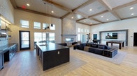 This single-floor space showcases an open-concept layout that seamlessly integrates a kitchen and lounge area under dramatically high ceilings. Exposed wood beams frame a coffered ceiling, adding warmth and architectural character to the expansive interior. Natural light floods the space through large windows positioned both at eye level and along the upper clerestory, creating a bright and airy atmosphere throughout the day.

The kitchen area features a large dark-toned island with a built-in sink, providing ample workspace and a striking visual contrast against the light hardwood flooring below. Sleek black pendant lights hang above the island, complementing the modern aesthetic. Adjacent to the kitchen, a stacked stone fireplace serves as a commanding focal point, rising toward the ceiling and anchoring the lounge area. The living space is furnished with generous sectional seating arranged around the fireplace, creating an inviting gathering environment. Light wood-toned walls and neutral finishes throughout tie the space together cohesively. A game area is visible in the background, suggesting this space is designed for communal use and entertainment.