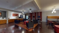 This single-floor commercial space is thoughtfully designed to accommodate both collaborative and private needs. The expansive main lounge serves as the heart of the space, featuring rich dark hardwood flooring, drop pendant lighting, and a tray ceiling that adds architectural depth. Seating arrangements include navy upholstered armchairs and bold red ottomans centered on a vibrant patterned area rug, creating an inviting yet professional atmosphere. Deep crimson accent walls with vertical light panels provide a striking visual contrast throughout the lounge.

A well-equipped beverage and service bar runs along one wall, outfitted with warm wood cabinetry, a coffee station, refrigerated display case, and ample counter space. Adjacent to the bar, a dedicated kitchen features white subway tile, stainless steel appliances, undermount sink, and pendant lighting above clean white countertops. Multiple wall-mounted televisions and bar-height workstations offer practical amenities throughout the lounge.

A formal conference room provides a more private setting, appointed with a large wood conference table, bold red chairs, built-in cabinetry, curated artwork, and a sliding barn door. Glass-paneled entry doors complete the polished, contemporary aesthetic of the overall space.