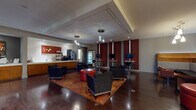 This single-floor commercial space is thoughtfully designed to accommodate both collaborative and private needs. The expansive main lounge serves as the heart of the space, featuring rich dark hardwood flooring, drop pendant lighting, and a tray ceiling that adds architectural depth. Seating arrangements include navy upholstered armchairs and bold red ottomans centered on a vibrant patterned area rug, creating an inviting yet professional atmosphere. Deep crimson accent walls with vertical light panels provide a striking visual contrast throughout the lounge.

A well-equipped beverage and service bar runs along one wall, outfitted with warm wood cabinetry, a coffee station, refrigerated display case, and ample counter space. Adjacent to the bar, a dedicated kitchen features white subway tile, stainless steel appliances, undermount sink, and pendant lighting above clean white countertops. Multiple wall-mounted televisions and bar-height workstations offer practical amenities throughout the lounge.

A formal conference room provides a more private setting, appointed with a large wood conference table, bold red chairs, built-in cabinetry, curated artwork, and a sliding barn door. Glass-paneled entry doors complete the polished, contemporary aesthetic of the overall space.