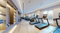 Located at 95 Anderson Street in Hackensack, New Jersey, this well-appointed fitness center offers a generously sized exercise room designed with both function and aesthetics in mind. The space features light hardwood-style flooring throughout, complemented by a coffered ceiling with elegant recessed and cove lighting that creates a bright, energizing atmosphere.

The cardio zone is equipped with multiple Matrix treadmills, stair climbers, and elliptical machines, all featuring integrated digital screens. Large windows line the perimeter, flooding the space with natural light and offering views of the surrounding Hackensack streetscape. Full-length mirrors along one wall visually expand the room and allow users to monitor their form during workouts.

The strength training area includes a comprehensive selection of Matrix cable machines, multi-functional weight stations, and a dedicated cycling bike, catering to a wide range of fitness goals. A central equipment rack houses stability balls and accessories, while warm wood-paneled accent walls and columns add a refined, upscale feel to the environment.

Practical amenities such as a water station and sanitizing dispenser are conveniently integrated near the entrance. The overall design balances a clean, modern aesthetic with a welcoming and motivating workout environment, making this fitness center a standout amenity in the heart of Hackensack.