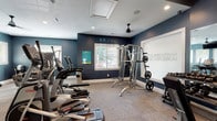 This fitness center offers a comprehensive workout environment designed to support a wide range of exercise routines. The space is finished with bold navy blue accent walls, white baseboards, and neutral carpeted flooring, creating a modern and motivating atmosphere. Multiple ceiling fans and recessed lighting ensure the area remains comfortable and well-lit throughout.

The cardio zone is stocked with elliptical trainers and treadmills, providing ample options for aerobic workouts. A dedicated strength training area houses a multi-function cable machine and a Smith machine with free weights, catering to both beginners and experienced lifters. A full dumbbell rack with a flat bench offers additional free weight training options, complemented by large wall-to-wall mirrors that enhance the sense of space and allow for proper form checks.

A functional fitness corner provides exercise balls, medicine balls, foam rollers, and yoga mats neatly organized on a dedicated rack. Natural light filters in through several windows fitted with blinds, connecting the interior to the surrounding greenery outside. A water dispenser and waste receptacle are conveniently placed within the space. An adjacent hallway with wood-style flooring leads to the entrance, completing this well-rounded and thoughtfully arranged fitness facility.