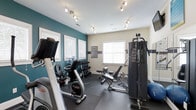 This fitness center offers a comprehensive workout environment designed to support a variety of exercise routines. The space is thoughtfully appointed with a mix of cardio and strength training equipment, including stationary bikes, a multi-function cable machine, a free weight rack with dumbbells, and an adjustable weight bench — providing everything needed for a full-body workout. The interior features a striking teal accent wall paired with neutral tones, creating an energizing yet focused atmosphere. Track lighting runs across the white ceiling, ensuring bright, even illumination throughout the space. Durable rubber flooring covers the workout area, providing both safety and comfort underfoot. Multiple windows dressed with white blinds allow ample natural light to fill the room while maintaining privacy. Fitness guidelines and rules are posted on the wall for member reference. The layout is efficiently organized to maximize usable floor space, allowing members to move freely between equipment. Overall, the fitness center presents a clean, modern, and functional environment well-suited for daily exercise.