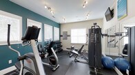 This fitness center offers a comprehensive workout environment designed to support a variety of exercise routines. The space is thoughtfully appointed with a mix of cardio and strength training equipment, including stationary bikes, a multi-function cable machine, a free weight rack with dumbbells, and an adjustable weight bench — providing everything needed for a full-body workout. The interior features a striking teal accent wall paired with neutral tones, creating an energizing yet focused atmosphere. Track lighting runs across the white ceiling, ensuring bright, even illumination throughout the space. Durable rubber flooring covers the workout area, providing both safety and comfort underfoot. Multiple windows dressed with white blinds allow ample natural light to fill the room while maintaining privacy. Fitness guidelines and rules are posted on the wall for member reference. The layout is efficiently organized to maximize usable floor space, allowing members to move freely between equipment. Overall, the fitness center presents a clean, modern, and functional environment well-suited for daily exercise.