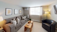 This single-floor residence offers a thoughtfully designed layout that balances comfort and functionality. The living room serves as the central gathering space, featuring plush wall-to-wall carpeting, neutral grey walls, and a large window dressed with sheer curtains that invite an abundance of natural light. The room is furnished with a spacious grey sofa, coordinating accent chairs, and rustic wooden coffee tables, creating a relaxed yet polished atmosphere. A wall-mounted flat-screen TV and a wall-unit air conditioner add modern convenience, while framed artwork and a decorative wall medallion lend personality to the space.

The bedroom is a serene retreat, appointed with soft carpet flooring, warm neutral tones, and a generously sized upholstered bed with a tufted headboard. Flanking bedside lamps cast a warm glow, and a large window with elegant draping allows for plenty of daylight while maintaining privacy. Baseboard heating is present throughout.

The galley-style kitchen is efficiently arranged with white cabinetry, granite-look countertops, and tile flooring. It is equipped with a full suite of white appliances, including a coil-top range, dishwasher, and refrigerator, making it a fully functional space for everyday cooking. The overall design of the residence emphasizes clean lines, neutral palettes, and comfortable, practical living.