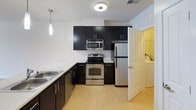 This single-floor residence is designed with a clean, contemporary aesthetic throughout. The kitchen serves as a functional centerpiece, outfitted with dark espresso cabinetry, stainless steel appliances including a refrigerator, electric range, and microwave, and light-colored countertops that extend into a peninsula with pendant lighting overhead. The hard surface flooring in the kitchen transitions seamlessly into carpeted living areas, which are bright and open with neutral cream-toned walls and white baseboards. The living space is generously sized, benefiting from natural light through multiple windows fitted with white blinds, and is equipped with a ceiling fan for added comfort. An additional carpeted room, suitable as a bedroom, also features windows that allow ample natural light to filter in. The bathroom is well-appointed with light tile flooring, a full soaking tub with shower combination, a toilet, and a dark-cabinetted vanity topped with a white sink. A linen closet provides convenient storage within the bathroom. Throughout the unit, the consistent use of neutral tones, modern fixtures, and practical finishes creates a cohesive and comfortable living environment.