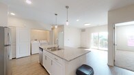 This well-designed single-floor unit offers a bright, open-concept layout that seamlessly connects the kitchen, dining, and living areas. The kitchen serves as the centerpiece, anchored by a generous island with a double stainless steel sink and granite countertops, complemented by pendant lighting overhead. White shaker-style cabinetry lines the walls, providing ample storage, while stainless steel appliances — including a refrigerator, electric range, and microwave — add a polished, modern finish. Light wood-style flooring runs throughout the main living areas, enhancing the clean, contemporary aesthetic. A dedicated in-unit laundry area with a full-size washer and dryer is conveniently accessible from the kitchen. Sliding glass doors in the living area lead to a private balcony, filling the space with natural light and offering outdoor views. The bedrooms feature soft carpeting, ceiling fans, and windows that bring in additional natural light, creating a comfortable retreat. The primary bathroom is spacious and well-appointed, featuring granite countertops, white cabinetry, a large mirror, a glass-enclosed shower, and direct access to a walk-in closet. A secondary bathroom mirrors the same refined finishes, maintaining a consistent design throughout the unit.