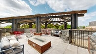 Located at 9 Municipal Dr in Fishers, Indiana, this outdoor rooftop terrace is a well-appointed communal space that combines functional design with an inviting atmosphere. The terrace is anchored by a striking wood-and-steel pergola structure that spans the main gathering area, providing partial shade while framing open sky views above. Large-format tile paving covers the floor throughout, lending a clean, modern aesthetic to the space.

At the center of the terrace sits a rectangular fire pit with a granite-topped surround and a wood base, serving as a natural focal point for the seating area. Comfortable lounge furniture, including cushioned sofas and wire chairs, is arranged around the fire pit to encourage relaxed gathering. Bistro-style tables and vibrant red folding chairs are positioned toward the perimeter, offering casual dining or conversational seating options.

Along the edges of the terrace, metal railings with built-in raised planter beds add a touch of greenery, featuring flowering plants and foliage that soften the urban setting. Views of the adjacent multi-story brick building with classic shuttered windows and balconies are visible from the terrace, adding architectural character to the surroundings. The open-air design floods the space with natural light, making it a welcoming destination for outdoor relaxation and social gatherings.
