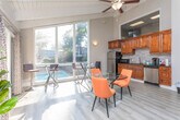This single-floor space showcases a mid-century modern aesthetic defined by its striking vaulted ceilings with exposed white-painted wooden beams that draw the eye upward and lend the interior a sense of generous height and openness. Light hardwood-style flooring runs throughout, unifying the various living and dining areas seamlessly.

The living area is a standout feature, oriented toward a wall of large windows that frame lush, tree-filled outdoor views and flood the space with natural light. A sliding door provides access to an adjoining balcony or deck. The room comfortably accommodates a tufted sofa, media console, and decorative greenery, giving it a relaxed yet curated feel.

Adjacent to the living area is a spacious dining zone anchored by a substantial dark wood table paired with a circular glass-top table and modern chairs. The kitchenette nearby features rich, dark wood cabinetry, a stone tile backsplash, stainless steel appliances, and ample counter space.

A staircase with warm wood treads and wrought iron railings rises from the main level, connecting to the floor above. Together, these elements create an inviting, light-filled space with a cohesive, nature-inspired atmosphere throughout.