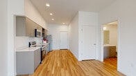 This contemporary single-floor unit offers a clean, modern aesthetic defined by crisp white walls, warm wood-tone luxury vinyl plank flooring, and an efficient, well-considered layout. The open-concept kitchen is fully equipped with stainless steel appliances — including a range, microwave, refrigerator, and dishwasher — complemented by sleek gray flat-front cabinetry, open shelving, and a white quartz countertop with an undermount sink. Recessed lighting illuminates the kitchen and main living corridor, creating a bright and functional environment. The living area extends naturally from the kitchen, offering an open, airy feel with a large window that invites natural light and provides views of the surrounding neighborhood. The two bedrooms are well-proportioned, each featuring large windows with neighborhood views and wall-unit climate control. One bedroom is finished with soft carpeting for added comfort, while a walk-in closet provides generous storage. The full bathroom is finished in a clean, minimalist style with wood-tone flooring, a gray vanity, and a full tub-shower combination. Throughout the unit, the cohesive design language, modern fixtures, and thoughtful storage solutions make this a practical and attractive living space.